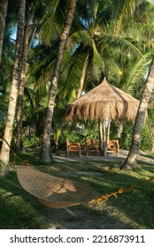 Tropical Backyard Scene With Lush Green Vegetation And Palm Trees Overhead, Supporting A Bamboo Wicker Hammock And Umbrella And Chair Table Set For Relaxation On Vacation Holiday Destination