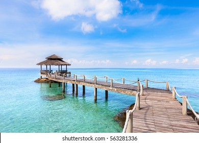 Tropical Beach Landscape With A Wooden Pier Or Jetty With Hut Above Transparent Turquoise Clear Sea Water And Blue Sunny Sky, Beautiful Vacation Destination