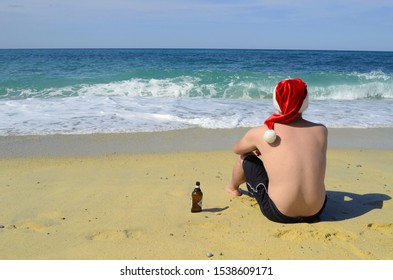 Tropical Christmas. A Man In A Hat Of Santa Claus Seets In The Beach And Drinks Beer On The Seashore.