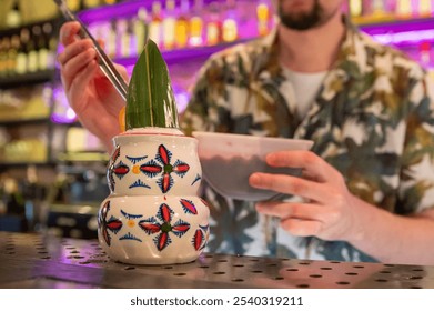 tropical cocktail in a decorative tiki mug garnished with a green leaf, set on a bar counter. The background features a blurred bartender and bottles, creating an inviting atmosphere for socializing. - Powered by Shutterstock