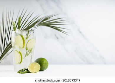 Tropical detox fresh citrus drink with ice, lime slices, green rosemary twig, silver straw, palm leaf on soft light white wood table and marble wall in sunlight.