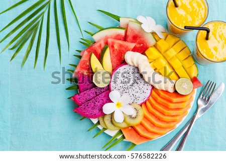 Tropical fruits assortment on a white plate with palm tree leaf with mango smoothie, textile blue background. Top view. Copy space