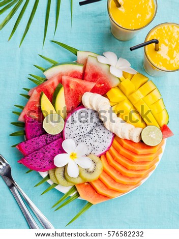 Tropical fruits assortment on a white plate with palm tree leaf with mango smoothie, textile blue background. Top view. Copy space