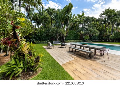 Tropical Garden With Long Table And Wooden Benches, Metal Armchairs With Fire Pit, Swimming Pool, Tiled Floor, Short Grass, Blue Sky With Clouds