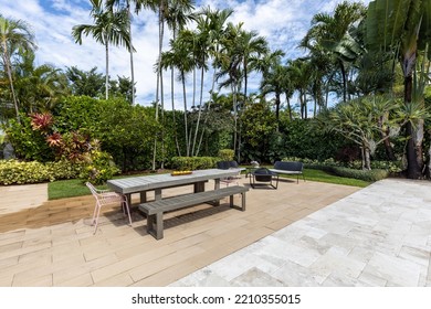 Tropical Garden With Long Table And Wooden Benches, Metal Armchairs With Fire Pit, Tiled Floor, Short Grass, Blue Sky With Clouds