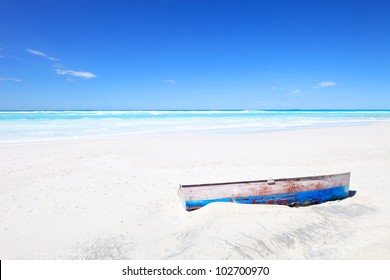 Tropical Seascape with an wooden, old and broken boat on white and sunny beach under a blue sky.