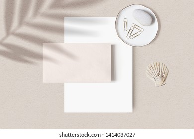 Tropical summer stationery mock-up scene. Blank business card, porcelain plate with stone, gold paper clips and sea shell, beige textured table background. Palm leaf shadow overlay. Flat lay, top view