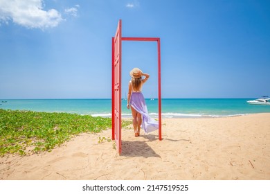 Tropical Travel Vacation. Traveler Woman Walks Out In Red Door On The Sandy Beach To Turquoise Sea, Toward Vacation And Relaxation In Sunny Asia. Carefree Girl Tourist In Swimwear In Asian Destination