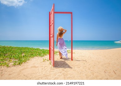 Tropical Travel Vacation. Traveler Woman Walks Out In Red Door On The Sandy Beach To Turquoise Sea, Toward Vacation And Relaxation In Sunny Asia. Carefree Girl Tourist In Swimwear In Asian Destination