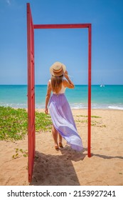 Tropical Travel Vacation. Traveler Woman Walks Out In Red Door On The Sandy Beach To Turquoise Sea, Toward Vacation And Relaxation In Sunny Asia. Carefree Girl Tourist In Swimwear In Asian Destination