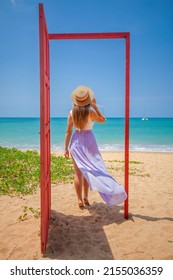 Tropical Travel Vacation. Traveler Woman Walks Out In Red Door On The Sandy Beach To Turquoise Sea, Toward Vacation And Relaxation In Sunny Asia. Carefree Girl Tourist In Swimwear In Asian Destination