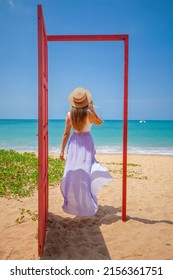 Tropical Travel Vacation. Traveler Woman Walks Out In Red Door On The Sandy Beach To Turquoise Sea, Toward Vacation And Relaxation In Sunny Asia. Carefree Girl Tourist In Swimwear In Asian Destination