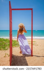 Tropical Travel Vacation. Traveler Woman Walks Out In Red Door On The Sandy Beach To Turquoise Sea, Toward Vacation And Relaxation In Sunny Asia. Carefree Girl Tourist In Swimwear In Asian Destination