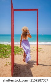 Tropical Travel Vacation. Traveler Woman Walks Out In Red Door On The Sandy Beach To Turquoise Sea, Toward Vacation And Relaxation In Sunny Asia. Carefree Girl Tourist In Swimwear In Asian Destination