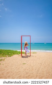 Tropical Travel Vacation. Traveler Woman Walks Out In Red Door On The Sandy Beach To Turquoise Sea, Toward Vacation And Relaxation In Sunny Asia. Carefree Girl Tourist In Swimwear In Asian Destination