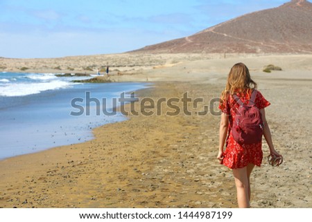 Tropical traveling. Young woman with red dress and backpack walking barefoot by sea beach enjoying landscape,