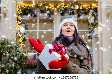 Troubled Brunette Woman With Unhappy Emotions Holding Present Box With Red Bow, Looking Up At Sky In Knitted Red Mittens Against Window With Garland Lights In Cold Winter Day. Snowfall Effect.