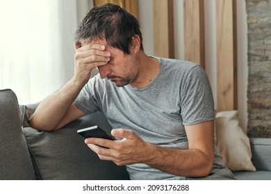 Troubled man receiving bed news in text message on mobile phone while sitting at living room sofa