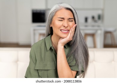 Troubled mature 50s woman with gray-hair feels strong toothache, holding cheek and suffering from strong dental pain sitting on sofa, middle-aged female undergoing tooth pain, enamel sensitivity - Powered by Shutterstock