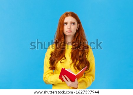 Troubled and pensive uneasy pretty redhead female, pouting and frowning looking away thoughtful, feeling sad and distressed, reading something bad in notebook, standing blue background