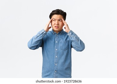 Troubled and uneasy asian man looking perplexed, having complicated situation. Guy with headache touching head and looking away, suffering painful migraine, standing white background - Shutterstock ID 1923728636