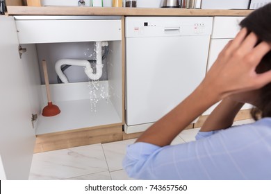 Troubled Woman Looking At Leaking Sink Pipe In Kitchen