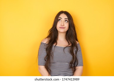 Troublesome puzzled Caucasian woman frowns face, bites lip, raises eyebrows, looks up, has problems, dressed in fashionable clothes, isolated over yellow background, has regret look.