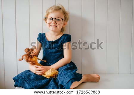 Trubschachen, Switzerland - December 16, 2019: Pretty caucasian little girl with short fair hair in blue dress holds her lovely barbie toy, sits in bright baby room and smiles