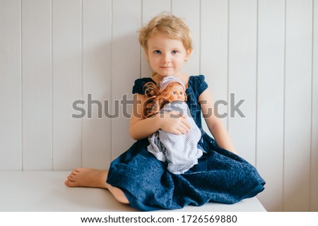 Trubschachen, Switzerland - December 16, 2019: Pretty caucasian little girl with short fair hair in blue dress holds her lovely barbie toy, sits in bright baby room and smiles