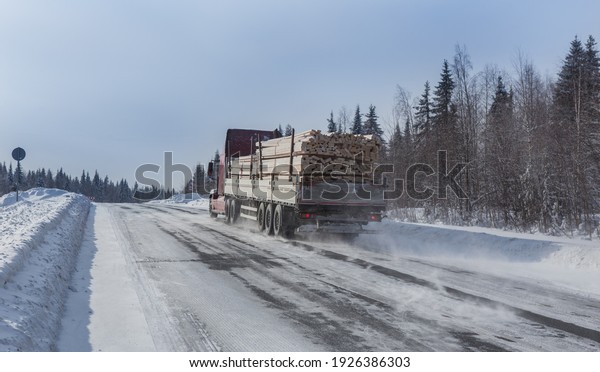 truck carries wooden planks along winter country\
road along the forest.