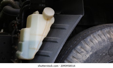 Truck Coolant Tank. The White Plastic Tank Has Coolant In The Engine Radiator System On A Dark Background And Rubber Wheels With Copy Space. Auto Parts Concept. Closeup And Focus On The Subject.