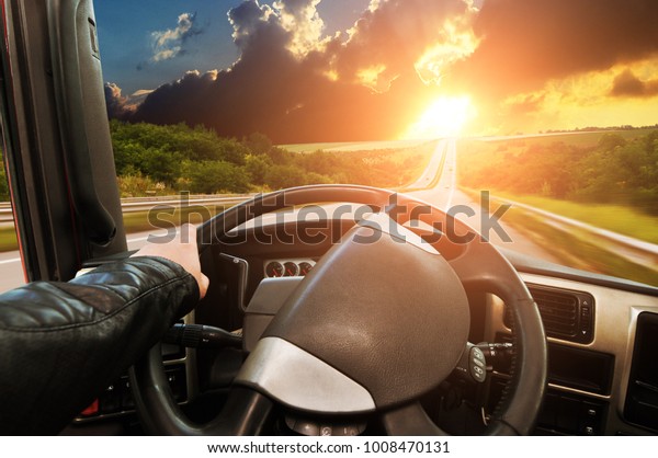 Truck\
dashboard with driver\'s hand on the steering wheel on the\
countryside road against night sky with\
sunset