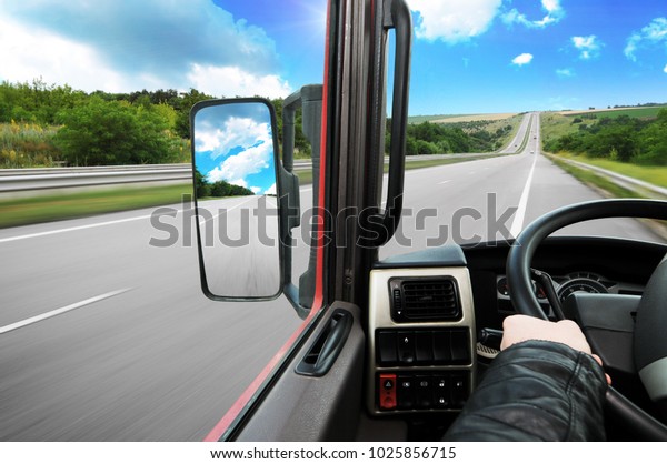 Truck dashboard with driver\'s hand on the steering\
wheel and side rear-view mirror on the countryside road against\
blue sky with clouds