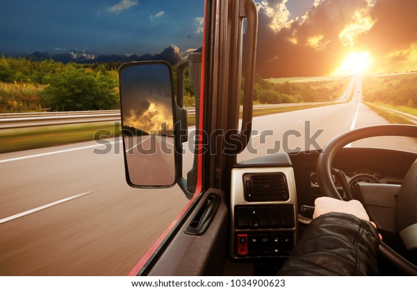 Truck dashboard with driver\'s hand on the steering\
wheel and side rear-view mirror on the countryside road against\
night sky with sunset