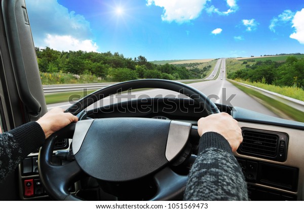 Truck dashboard with driver\'s hand on the steering\
wheel on the countryside road in motion against blue sky with\
sun