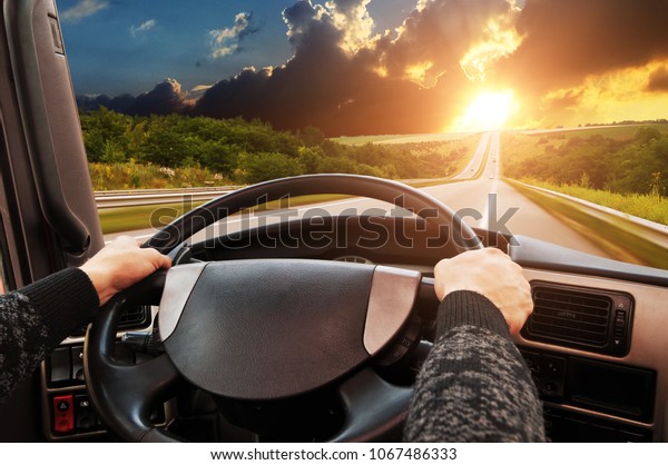 Truck dashboard with driver\'s hand on the steering\
wheel on the countryside road in motion against night sky with\
sunset
