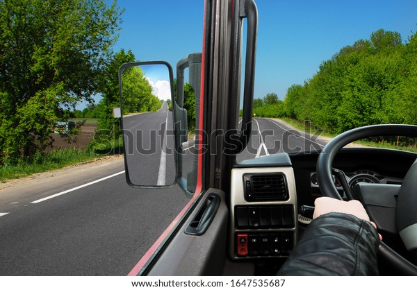 Truck dashboard with driver\'s hand on the steering\
wheel and side rear-view mirror on the countryside road against\
blue sky