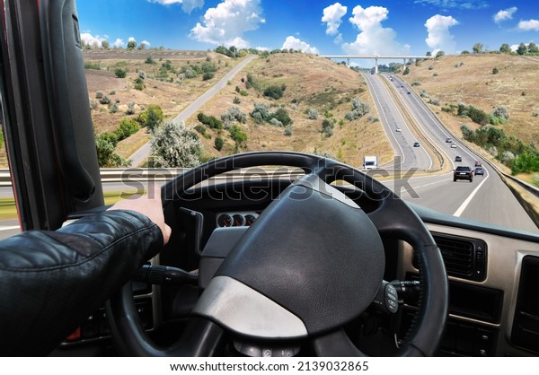 Truck dashboard with driver\'s hand on a black\
steering wheel against a countryside road in motion with cars and a\
blue sky with clouds