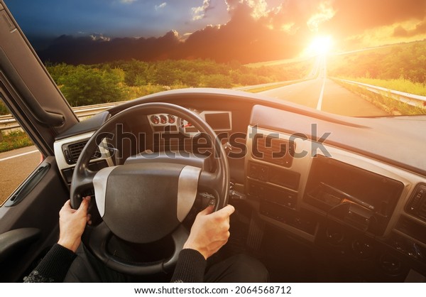 Truck\
dashboard with driver\'s hands on the steering wheel against\
countryside road with trees and a sky with a\
sunset
