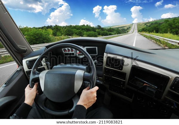 Truck dashboard with driver\'s hands on the\
steering wheel against countryside road with trees and a blue sky\
with clouds