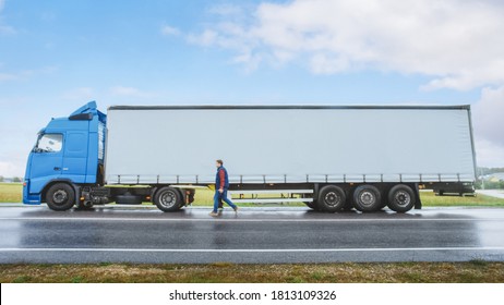 Truck Driver Crosses The Road In The Rural Area And Gets Into His Blue Long Haul Semi-Truck With Cargo Trailer Attached. Logistics Company Moving Goods Across Countrie Continent. Side View Shot