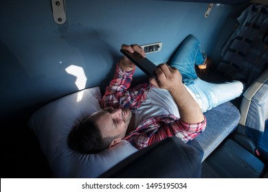 Truck Driver Lying On Bed In His Truck Cabin Behind Driver Seat And Holding Tablet Chatting With His Family. Trucker Lifestyle Missing Family.