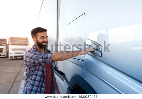 Truck driver occupation. Young bearded trucker\
opening truck vehicle door to enter the cabin and start driving.\
Transportation services.