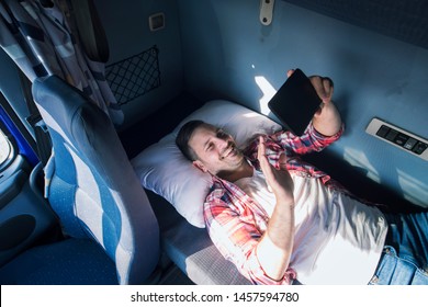 Truck Driver Separated From His Family Lying On The Bad Of His Truck Cabin And Waving To His Wife And Children Via Tablet Computer. Trucker Lifestyle Missing Family.