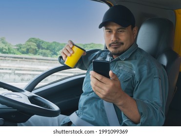 Truck Driver Video Call On Smartphone In Cockpit. Trucker Asian Young Man Beard Parked For Coffee Break After Long Drive. Worker Male Driving Semi-truck Transport Logistics Express Delivery Services.