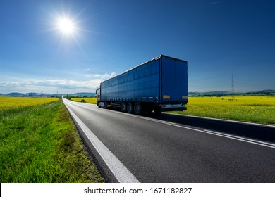 Truck driving on the asphalt road between the yellow flowering rapeseed fields under radiant sun in the rural landscape