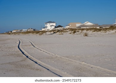 a truck leaves tire prints in the beach sand close to shore with beach houses in the background on a sunny day - Powered by Shutterstock