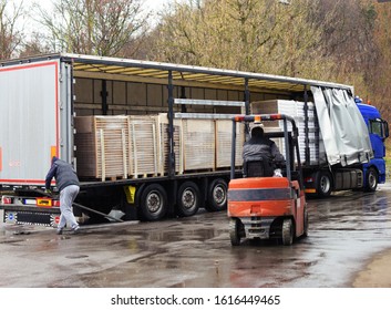 A truck is loaded with goods wrapped on pallets, a trucker is making a move while lowering the side board and the forklift is moving while approaching the goods to unload it while raining.