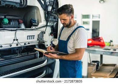 Truck Mechanic Writing Notes In Notebook During Working In Modern Workshop. Young Concentrated Caucasian Bearded Man.Truck Service, Repair. Garage Interior With Tools And Equipment