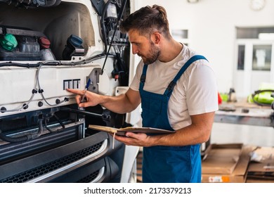 Truck Mechanic Writing Notes In Notebook During Working In Modern Workshop. Young Concentrated Caucasian Bearded Man.Truck Service, Repair. Garage Interior With Tools And Equipment